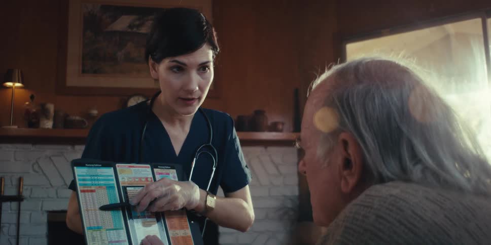 A healthcare professional shows a medical chart to an elderly man in a cozy room with a fireplace in the background
