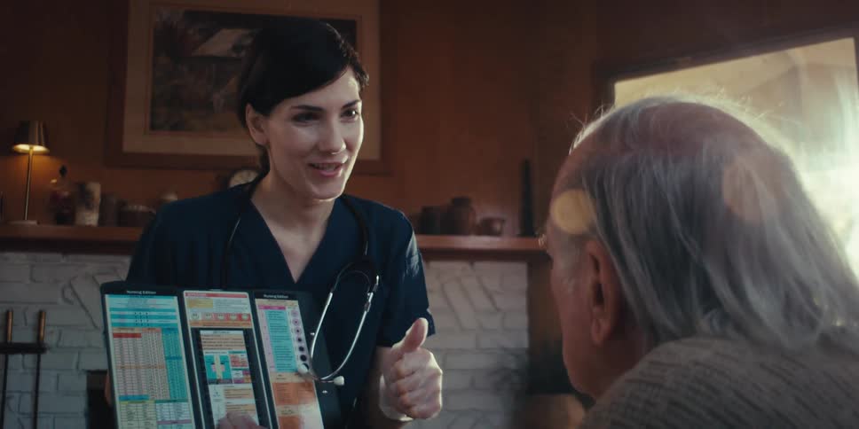 A healthcare worker in dark scrubs and a stethoscope is showing medical information on a chart to an elderly patient with gray hair