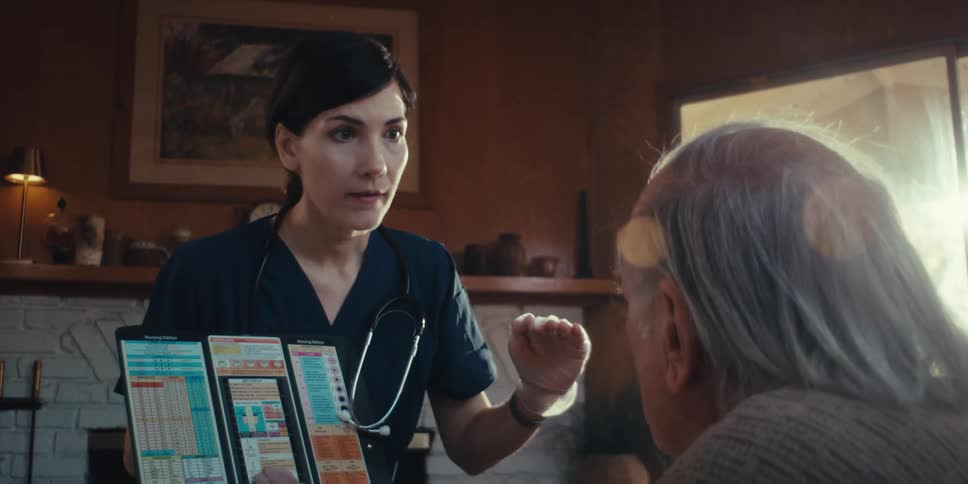 Female healthcare professional references a chart during a conversation with an elderly patient in his home