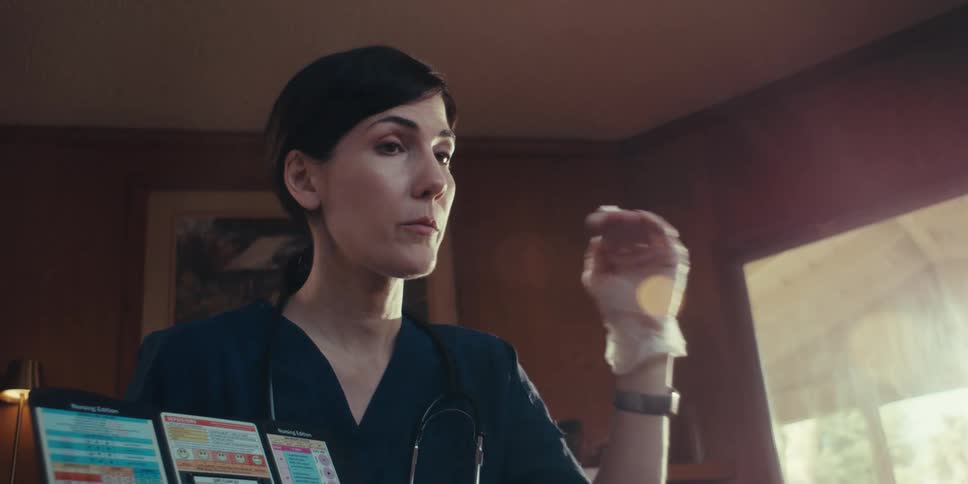 A healthcare professional wearing a dark scrub top gloves and a stethoscope is gesturing with one hand while looking upwards with medical charts visible in the foreground