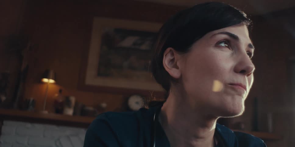 A health professional with dark hair pulled back wearing a dark scrub looking upwards and slightly to the right with a thoughtful expression in an indoor setting with blurred background details including a lamp and artwork