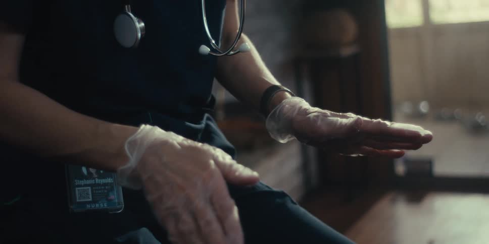 A closeup shows gloved hands of a nurse with a stethoscope around her neck gesturing while explaining in a home setting