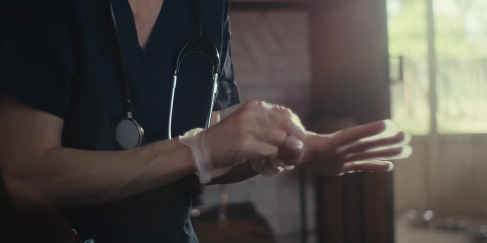 A healthcare professional wearing scrubs and a stethoscope carefully puts on disposable medical gloves
