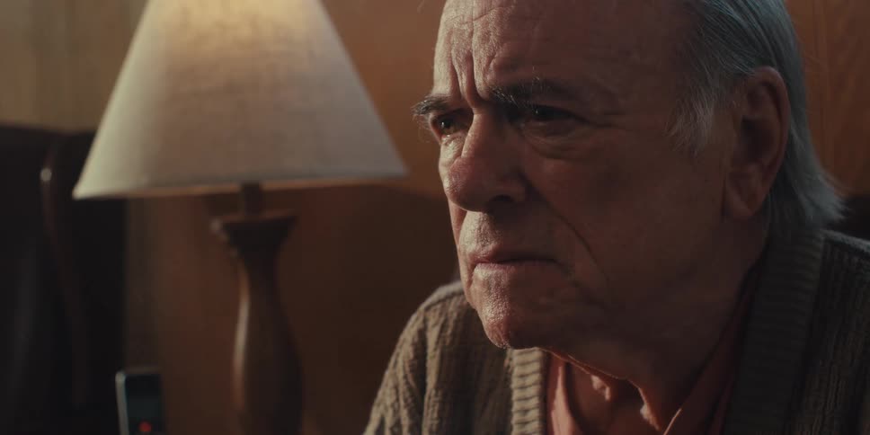 The somber expression of an older man with thinning white hair wearing a brown cardigan as he looks slightly to his right in a dimly lit room with a lamp visible in the background