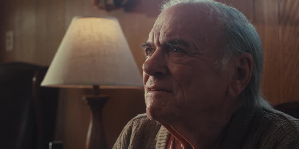 An elderly man with grey hair and teary eyes is looking slightly upwards and to the left listening emotionally with a softly illuminated lamp visible in the blurred background