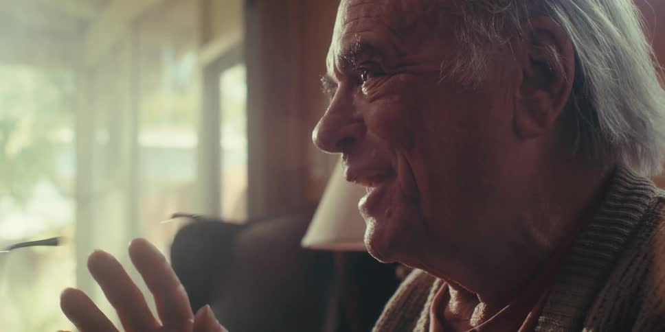 A closeup of an elderly man sitting on a couch at home smiling and talking to someone on a bright sunny day