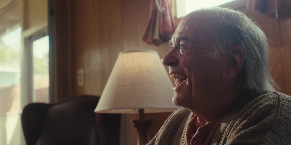 An elderly man sits at home removes his glasses and exhales with a sigh of relief while talking to someone nearby