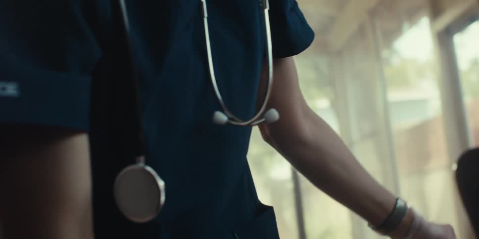 A health professional wearing a dark blue scrub top with a silver stethoscope hanging around their neck against a blurred background