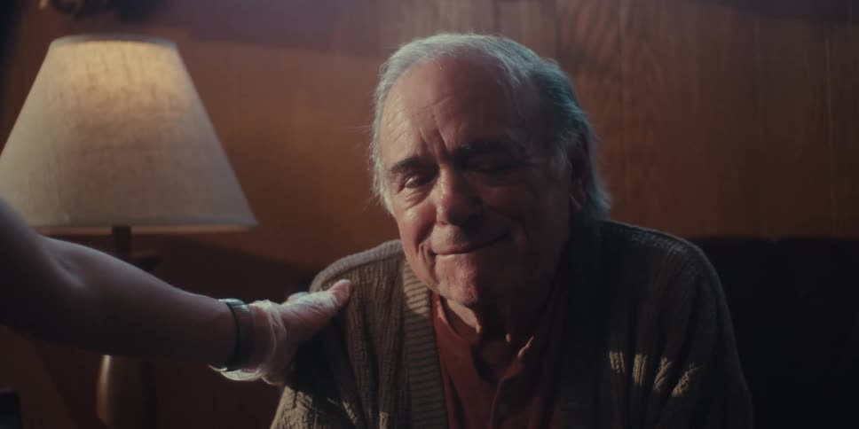 An older man with thinning white hair wearing a brown cardigan looking slightly to his right with a tearful expression as a hand wearing a white glove rests reassuringly on his shoulder in a dimly lit room with a lamp visible in the background