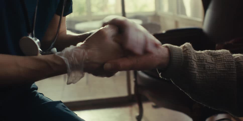 A healthcare professional wearing gloves and a stethoscope is holding the hand of an older person wearing a brown sweater