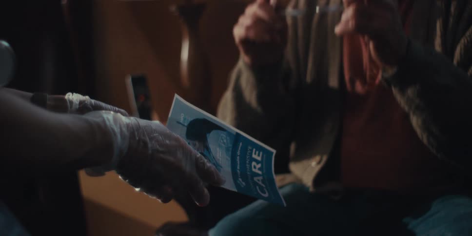 A nurse explains the contents of a brochure to an elderly man seated on a couch at home