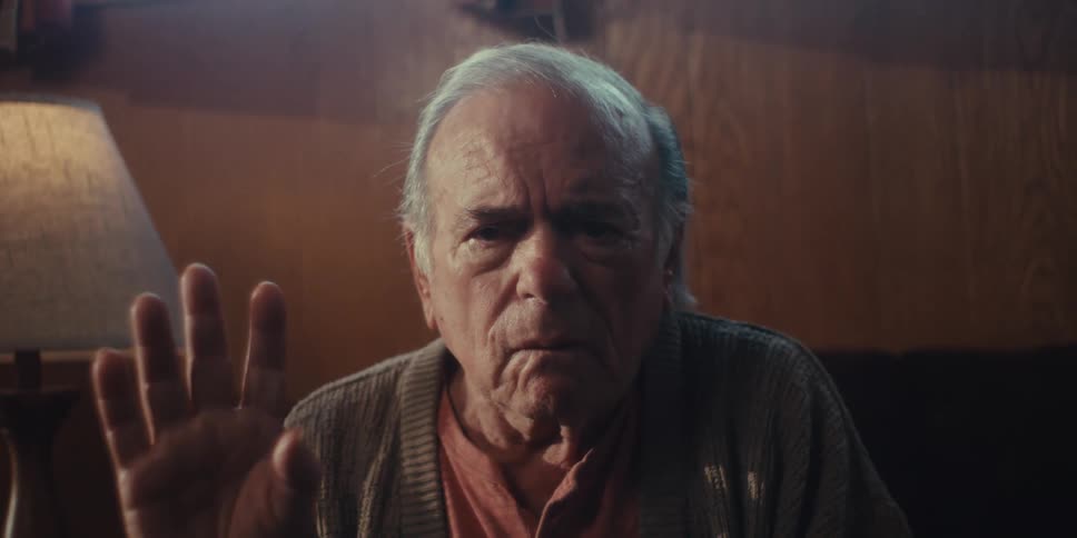 An elderly man sits at home with a sorrowful expression making a denying gesture with his hand and head