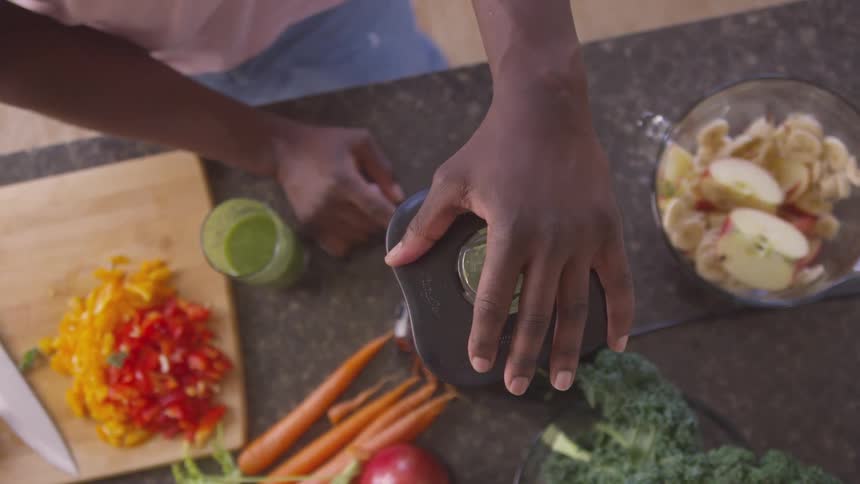 A black person is blending a green smoothie with chopped fruits and vegetables