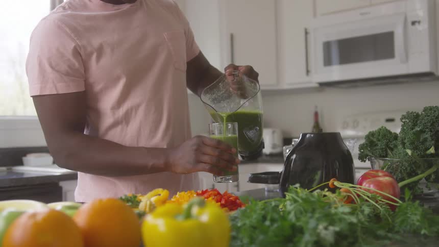 A man in a pink shirt pours a green smoothie from a pitcher into a glass surrounded by fresh fruits and vegetables in a bright kitchen