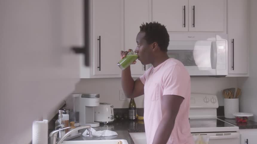 In a brightly lit kitchen with white cabinets and black countertops a man wearing a light pink t shirt is standing and drinking from a clear glass filled with a green liquid