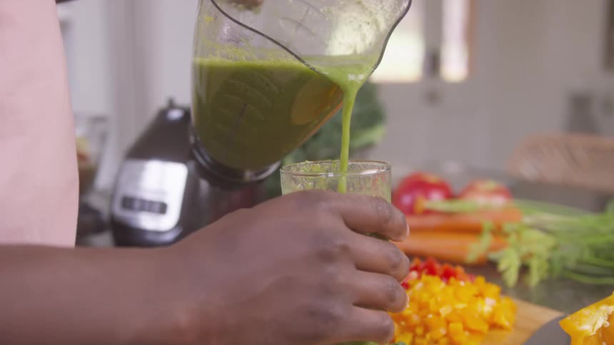 A closeup of a person pouring vegetable juice from a blender into a glass beside a table with chopped vegetables
