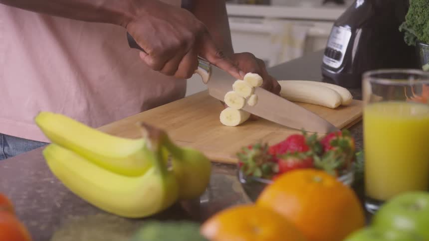 A man slicing bananas on a cutting board amidst various fruits like whole bananas strawberries oranges and a green apple on a kitchen counter with a blender and a glass of yellow drink also present