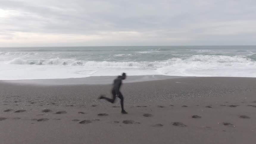 A man wearing a dark full body suit is running on a gray sandy beach next to a wavy ocean under a cloudy sky leaving footprints in the sand