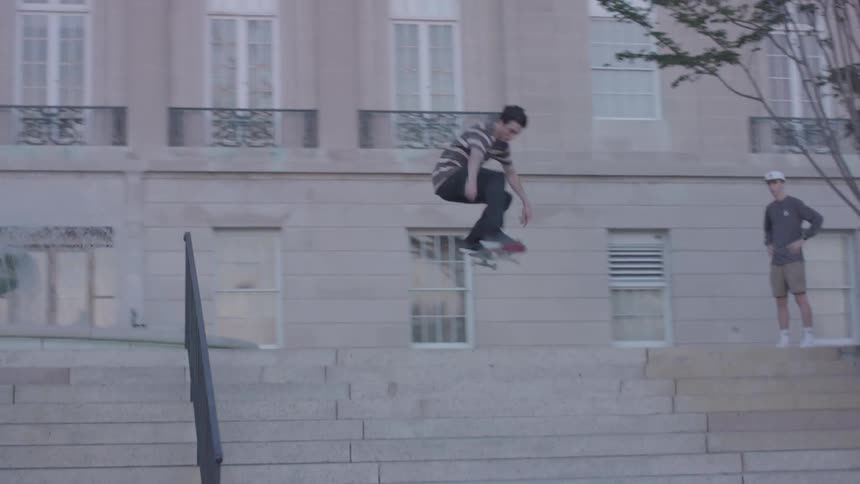 A skateboarder executes an aerial trick over stairs outside a building while two observers stand in the background