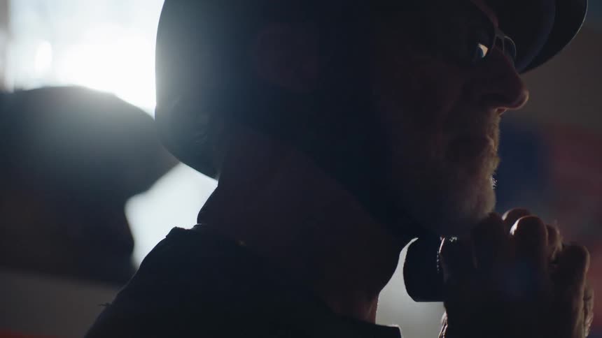 A closeup of the profile of a man in a dimly lit room as he puts on a motorcycle helmet