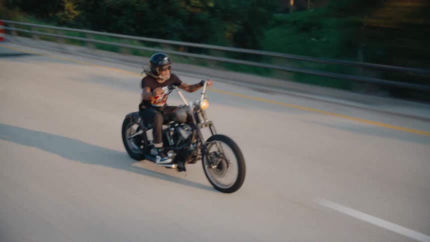 A man with long dreadlocks wearing a black helmet sunglasses a black t shirt and dark pants is riding a black and silver chopper motorcycle on a highway