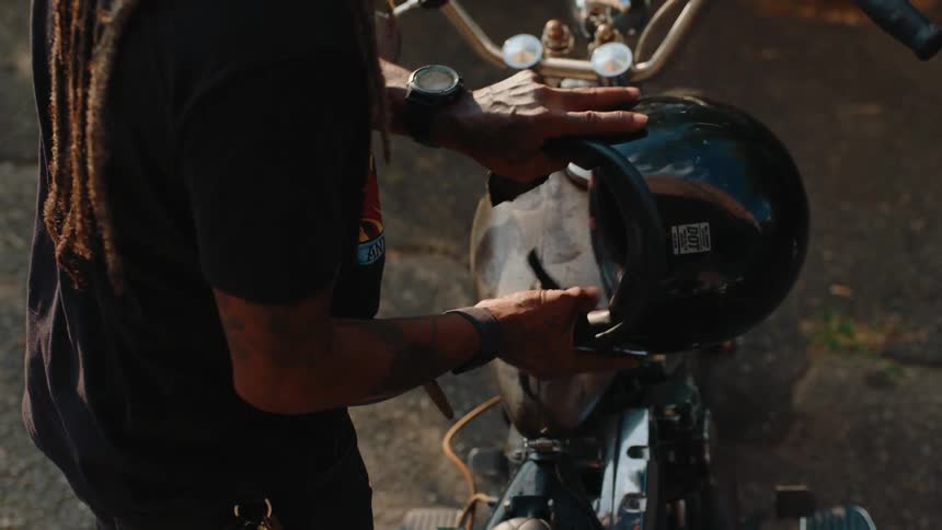 A man with long dreadlocks wearing a dark tshirt and a watch holding a black helmet near the handlebars of a vintagestyle motorcycle
