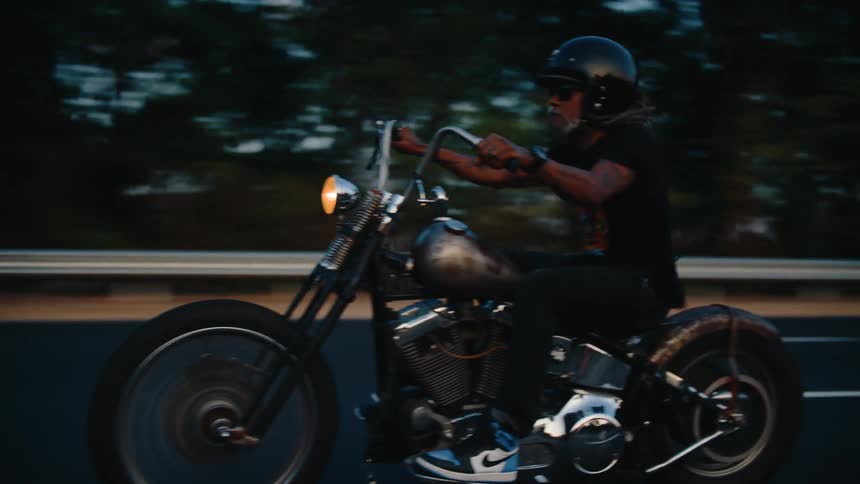 A man with long hair and a beard rides a motorcycle on a highway with a cityscape visible in the background