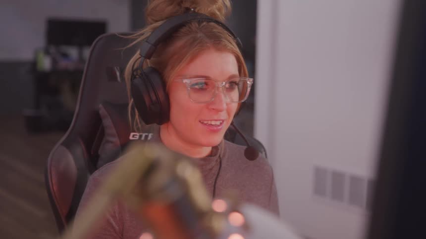 A woman sits in a gaming chair wearing headphones yelling with excitement while having fun playing a video game