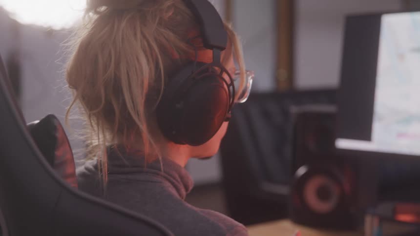 A woman sits in a gaming chair puts on headphones and prepares to play a video game using a controller