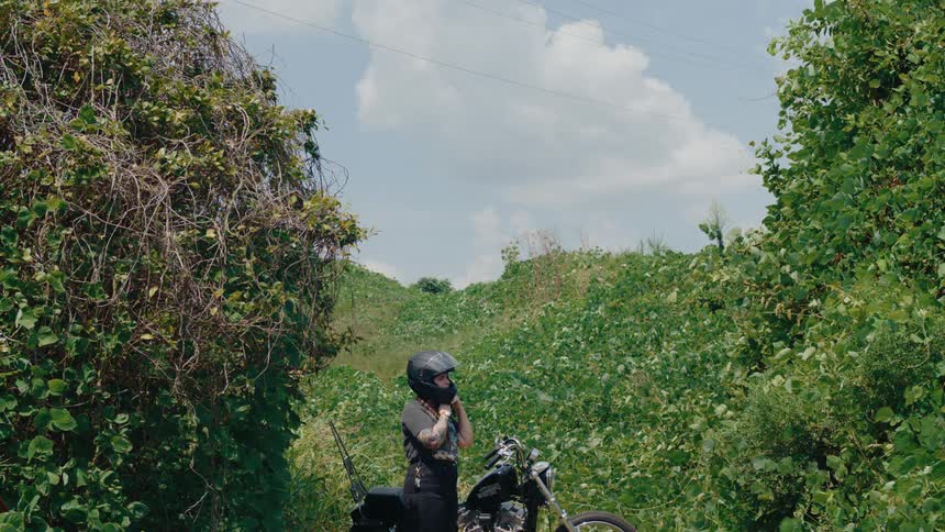 A woman adjusts their helmet while standing beside a motorcycle in a lush green area