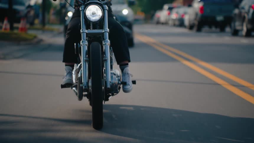 A front view of a motorcycle rider heading towards the viewer on an older style motorcycle followed by a car on a well maintained street while in the background a line of cars line up at a red light