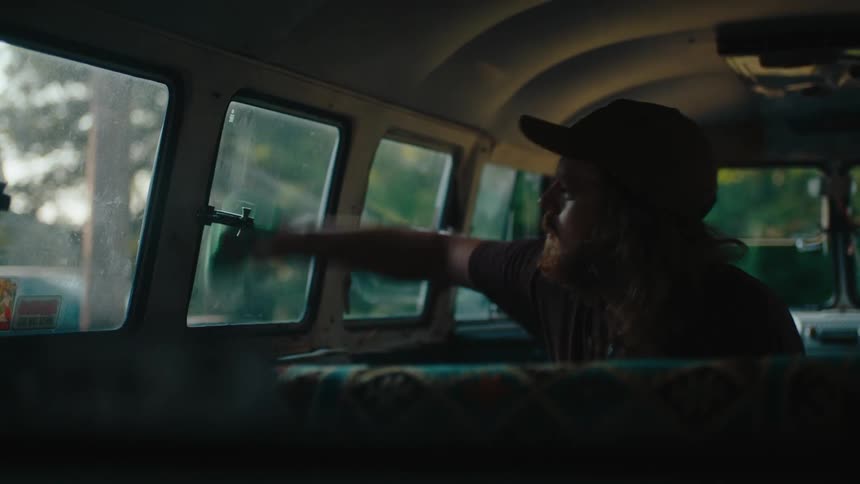 A person cleans the windows inside a vintage van using a cloth with sunlight streaming through surrounding trees
