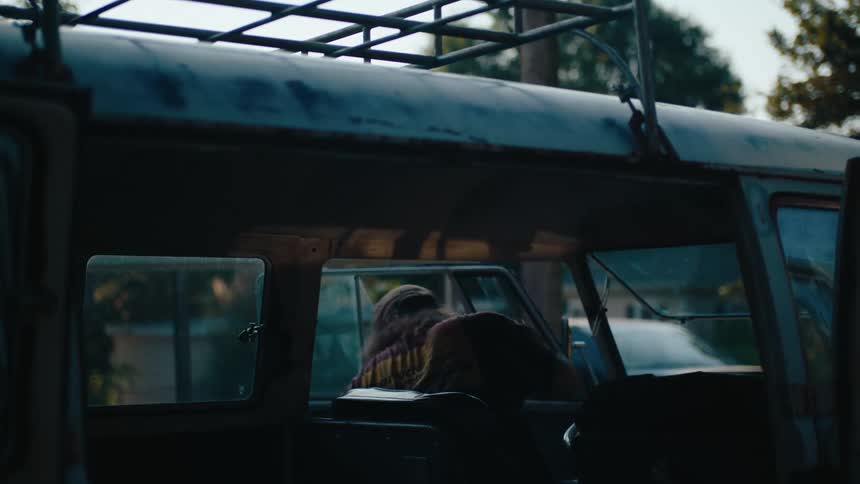 A person sits inside an old van with a roof rack surrounded by dim light as sunlight filters through the windows