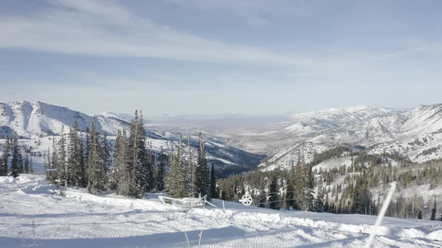 Several individuals operate near snow grooming machinery in a vast snowy mountain terrain surrounded by pine trees and distant peaks under clear skies