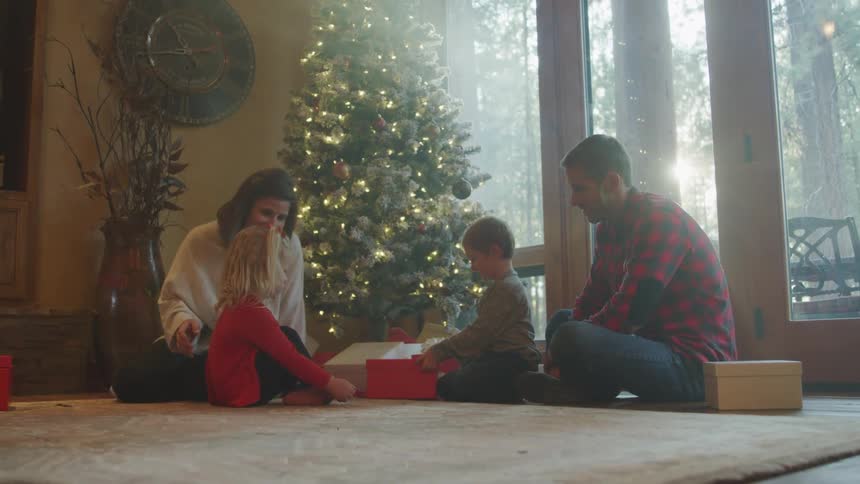 Family sitting on carpet next to christmas tree with two young children opening presents