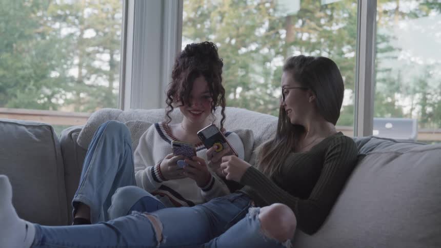 Two young women sitting together in the living room smile while enjoying their smartphones