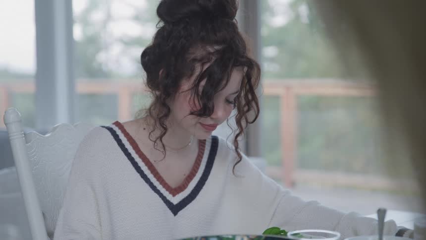 A teenage girl in a white sweater with a striped neckline is seated at a table praying with her family before eating