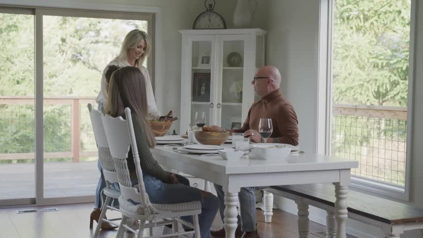 A family of four prepare to enjoy a meal at a dining table with a panoramic view of a lush green forest through a large window
