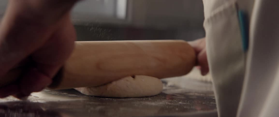 Hands holding a rolling pin and kneading bread dough on a metal surface