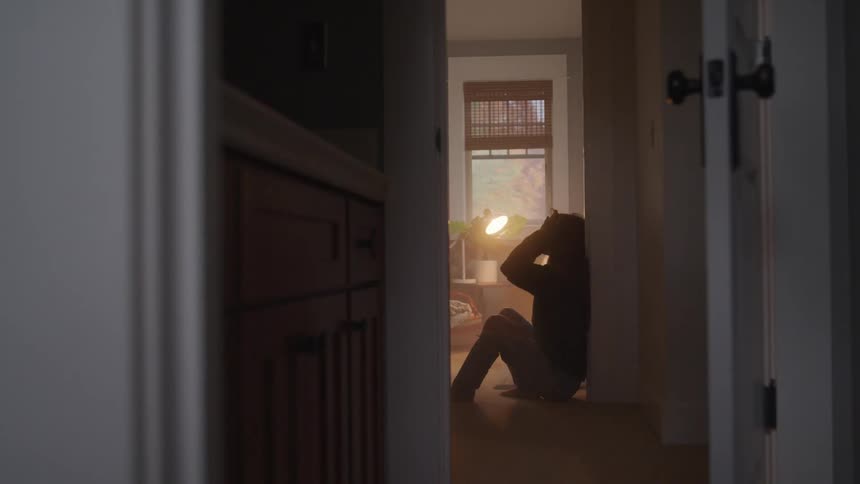 Viewed down a dimly lit hallway a silhouetted person slowly sinks to the floor against a wall holding their head in apparent distress
