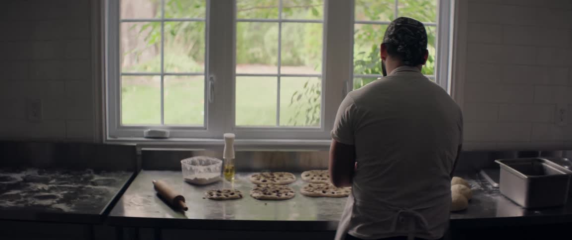 A person in a white shirt and apron prepares bread on a stainless steel counter with a garden visible through a large window
