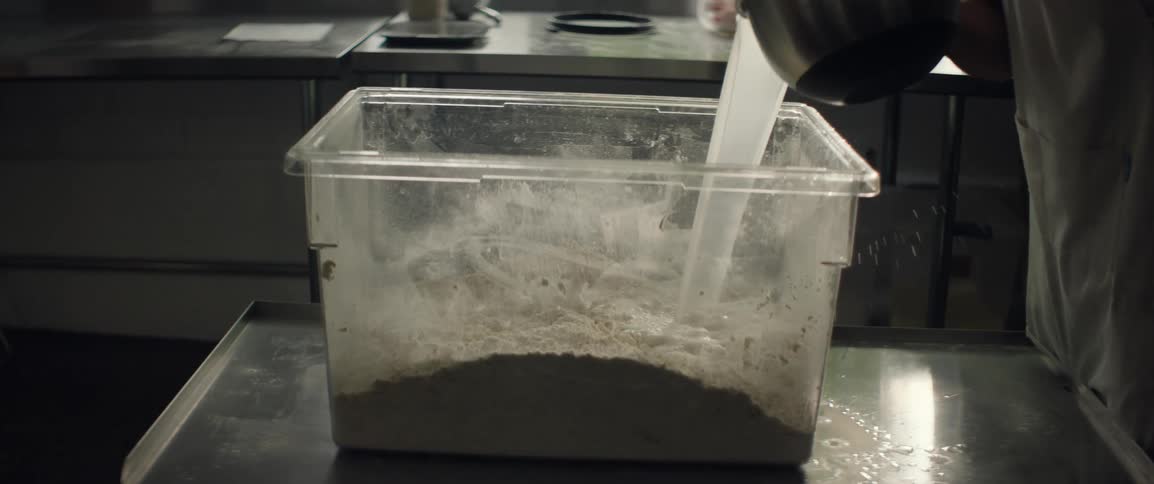 A baker pours water into a large container containing flour to make bread dough