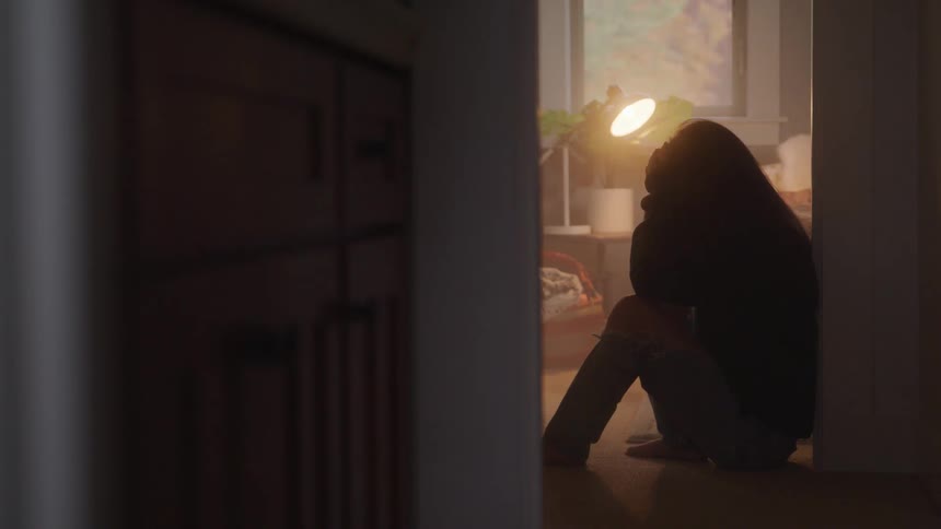 A teenage girl sits in a dimly lit room holding her head in both hands conveying sadness or distress