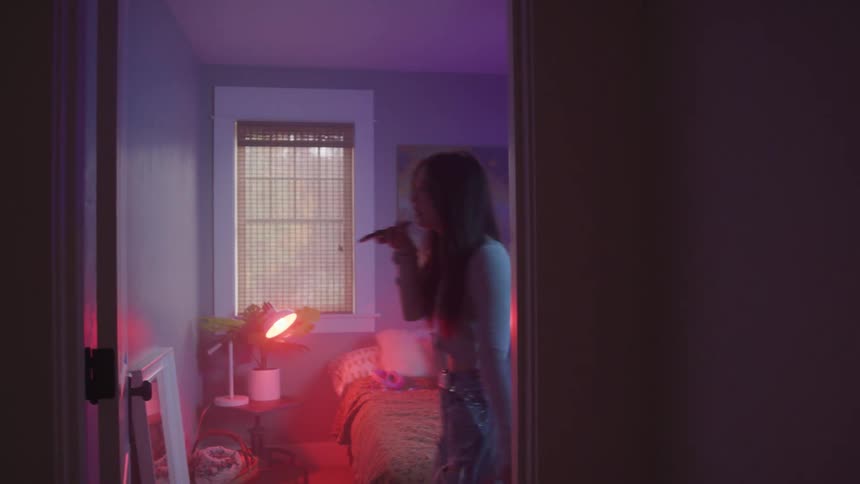 A teenage girl paces a dimly lit bedroom bathed in red and purple light while speaking on a smartphone
