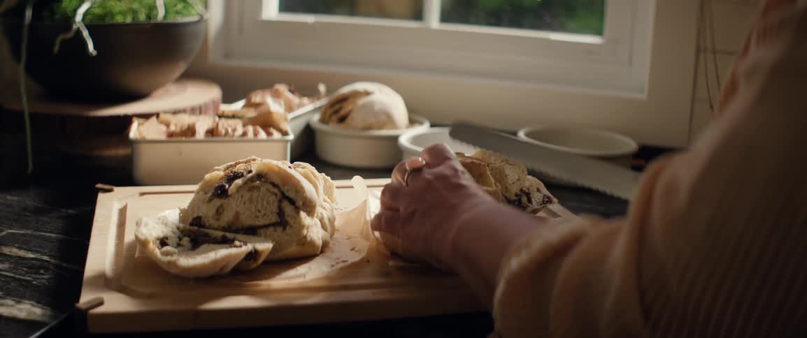 A person uses a large knife to carefully slice a freshly baked loaf of bread on a wooden cutting board in a sunlit kitchen