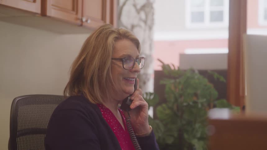 A woman with glasses talks pleasantly on a landline phone while sitting at her office desk