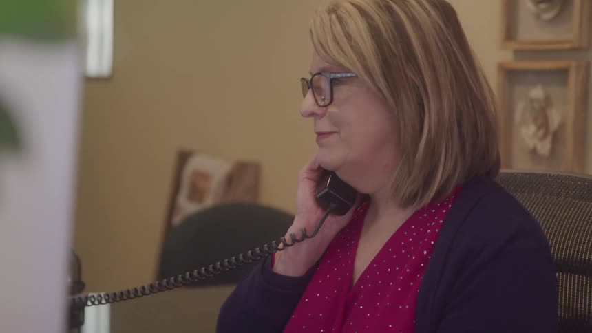 A woman wearing glasses speaks on a corded landline phone while seated behind a reception desk in an office hallway surrounded by framed art and softly lit walls