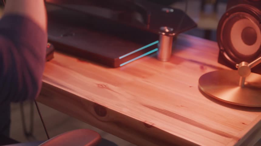 A person begins a gaming session by picking up a headset and preparing to play at a wooden desk setup with a game controller speaker and rgblit monitor stand