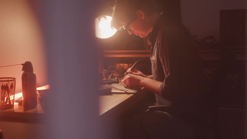 A person carefully assembles an electronic device at a desk illuminated by a bright lamp with various tools and components spread around