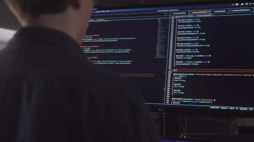 A man stands in front of a large monitor editing code with lines of programming text visible on the screen